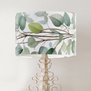 May include: A white lampshade featuring a watercolour design of green leaves and branches. The lampshade is on a white metal lamp base with an ornate design.