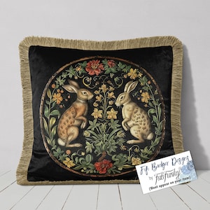 May include: Black square throw pillow with a gold fringe. The pillow features a circular design with two rabbits surrounded by flowers in a garden setting. The rabbits are facing each other.