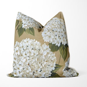 Puede incluir: Una almohada beige con un patrón floral blanco. La almohada presenta una gran flor de hortensia blanca con hojas verdes.