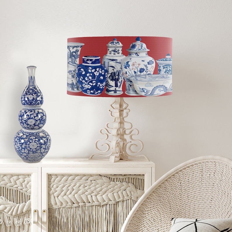May include: A white table lamp with a red lampshade featuring a pattern of blue and white porcelain vases. The lampshade has a white trim around the top and bottom. The lamp base is white and has a decorative design.