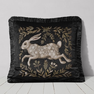 May include: Black velvet pillow with a white rabbit running through a floral design. The rabbit is covered in small white flowers and the background is a dark green.