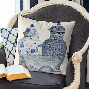 Op de afbeelding: Een wit kussen met een blauw en wit ontwerp van twee Chinese gembervazen en een kom met een drakenontwerp. Het kussen ligt op een grijze fauteuil.