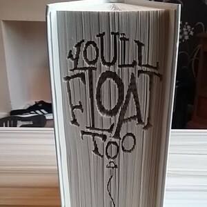 May include: A white book with the words "You'll Float Too" folded into the pages. The book is standing upright on a light wood surface.