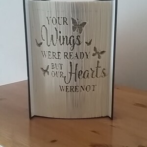 May include: A book with a black cover and a white paper interior. The pages are folded to create a 3D design of a butterfly with the text "Your Wings Were Ready But Our Hearts Were Not".