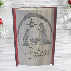 May include: A book art sculpture with a red spine and a gray cover. The pages are folded to create a three-dimensional image of the nativity scene with a star above the figures. The word "Believe" is written below the scene.