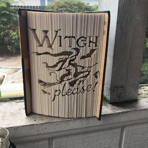 Op de afbeelding: Een boek met een gevouwen papieren ontwerp van een heks die op een bezem vliegt met de tekst "Witch please!"