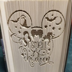 Op de afbeelding: Een gevouwen boek met een Halloween-ontwerp. Het ontwerp toont Mickey Mouse oren met vleermuizen, sterren en een heksenhoed. De tekst "I smell children" is in de pagina's van het boek gesneden.