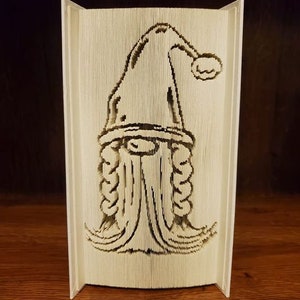 May include: A carved book sculpture featuring a gnome design. The gnome has a pointed hat with a pom-pom, a long beard, and braided hair. The sculpture is made from the pages of a book, with the design cut into the pages.