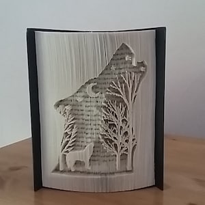 May include: A book sculpture depicting a wolf howling at the moon, with trees and a standing wolf carved into the pages. The artwork is set within a black frame, showcasing the intricate design.