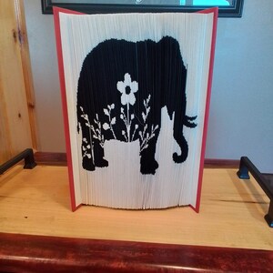 Peut inclure: Une silhouette noire d'un éléphant avec un pot de fleurs et des fleurs sur le dos. L'éléphant est fait de pages pliées d'un livre. Le livre est rouge et blanc.
