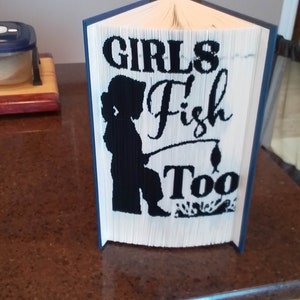 May include: A folded book sculpture with a blue spine and white pages. The front of the book features the words "GIRLS Fish Too" and a silhouette of a person fishing. The book is displayed on a brown countertop.