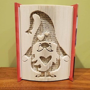 May include: A book art sculpture of a gnome with a heart on its chest. The gnome is made from the pages of a book that has been folded and cut to create the shape. The sculpture is standing on a wooden surface.
