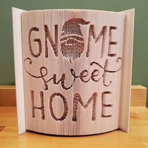 May include: A white book with the words "Gnome Sweet Home" cut out of the pages. The book is folded into a three-dimensional shape, creating a gnome figure in the center.