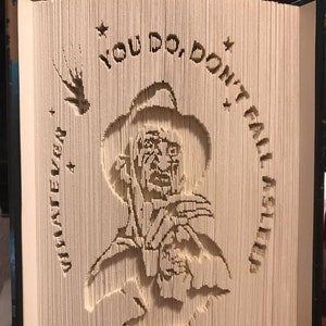 Puede incluir: Una escultura de libro de arte de Freddy Krueger de la película "A Nightmare on Elm Street". La escultura está hecha de papel y está doblada en un diseño 3D. La escultura es de la cabeza y los hombros de Freddy Krueger, y él está sosteniendo su guante característico con cuchillas. La escultura está rodeada por un círculo de estrellas y el texto "You do, don't fall asleep".