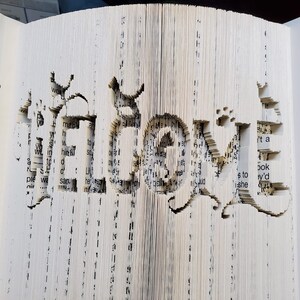 Peut inclure: Un livre avec le mot "Hello" découpé dans les pages. Les pages sont pliées et superposées pour créer un effet 3D.