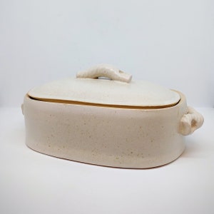 May include: A white ceramic oval baking dish with a lid. The lid has a unique, sculpted handle. The dish has a speckled glaze.