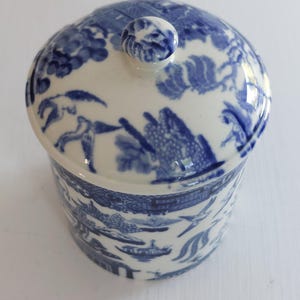 May include: A blue and white ceramic jar with a lid. The jar and lid feature a detailed landscape design. The lid has a small knob on top. The jar is round and has a matching design around the sides.