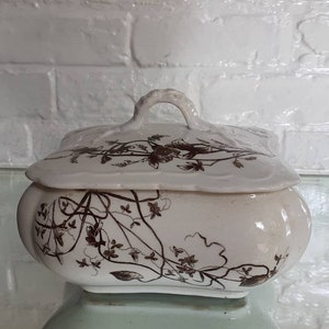 May include: Cream-colored ceramic lidded dish with a brown floral pattern. The dish has a rounded base and a lid with a handle. The floral design features vines and flowers. The background is a white brick wall.