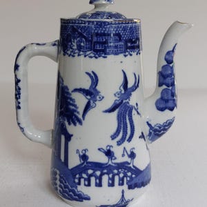 May include: A white and blue ceramic teapot with a lid. The teapot features a handle and spout, and is decorated with a blue and white landscape design. The design includes birds, bridges, and buildings. The teapot is a classic piece of kitchenware.