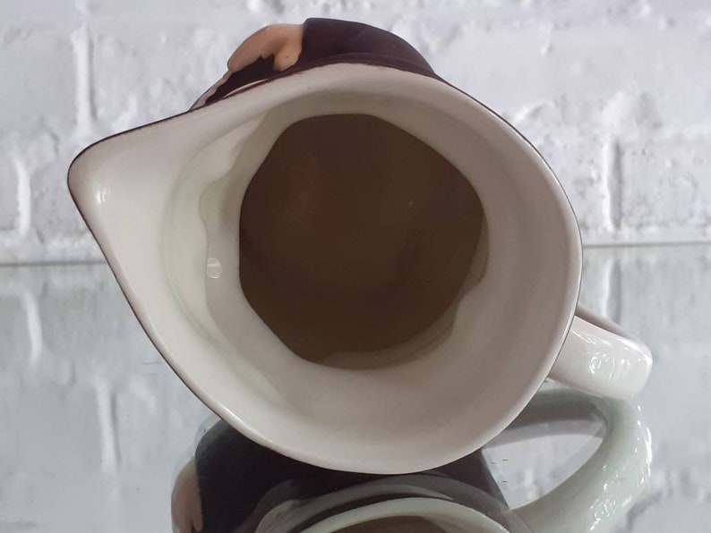 May include: A white ceramic pitcher with a brown interior and a dark brown rim. The pitcher has a handle and a spout. The image is taken from above, showing the inside of the pitcher.