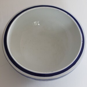 May include: A white ceramic bowl with a dark blue stripe around the rim. The bowl has a simple, minimalist design, suitable for serving food or as a decorative piece. The interior is plain white.