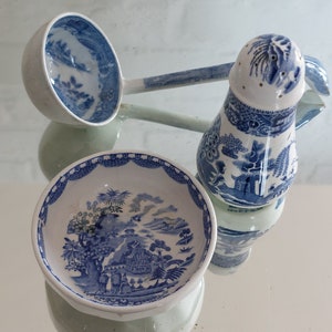 May include: A collection of blue and white ceramic tableware, including a ladle, a salt shaker, and a small bowl. The pieces feature a classic landscape design. The items are arranged on a reflective surface.