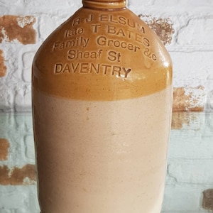 May include: A vintage stoneware jug with a tan and cream color scheme. The jug features embossed text that reads "R J Elsum late T Bates Family Grocer & c Sheaf St DAVENTRY". The jug has a rounded body and a narrow neck.