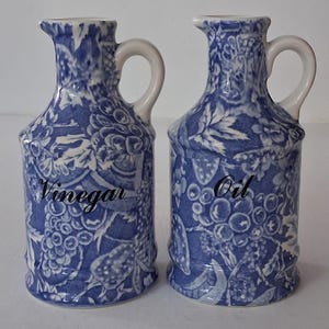May include: Two matching blue and white ceramic cruet bottles with handles. One bottle is labeled "Vinegar" and the other "Oil". Both bottles have a floral and fruit pattern.