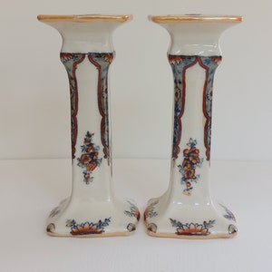 May include: Pair of white ceramic candlesticks with floral and geometric patterns in blue, orange, and brown. The candlesticks have a square base and a flared top, with a height of approximately 7 inches.
