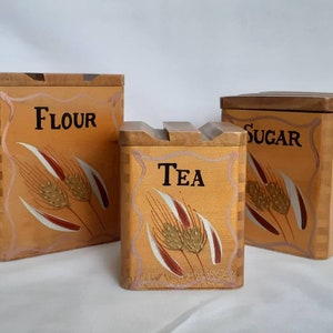 Puede incluir: Juego de tres botes de cocina de madera etiquetados como "FLOUR", "TEA" y "SUGAR". Cada bote tiene un dise&ntilde;o de espiga de trigo en tonos dorados, blancos y burdeos. Los botes son cuadrados con tapas de madera.