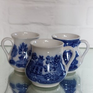 May include: Three small, white and blue ceramic mugs with a classic blue and white landscape design. Each mug has a curved handle and a rounded body. The mugs are displayed on a reflective surface.