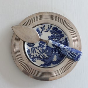 May include: A silver pie server rests on a small blue and white plate, atop a larger silver plate. The small plate features a blue willow pattern. The server has a silver blade and a blue and white patterned handle. The plates are on a white surface.