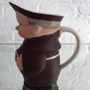May include: A ceramic pitcher shaped like a cartoon monk, featuring a brown robe, a white collar, and a white handle. The pitcher has a light skin tone and a brown hat. The base of the pitcher is green.