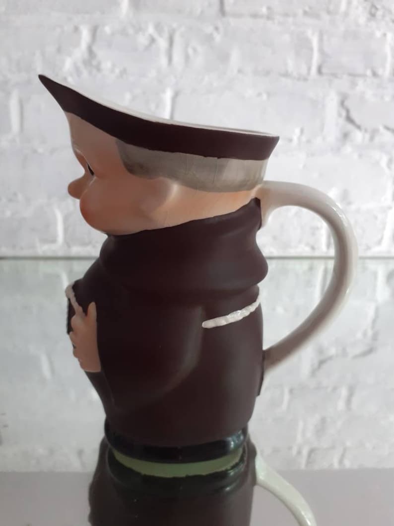May include: A ceramic pitcher shaped like a cartoon monk, featuring a brown robe, a white collar, and a white handle. The pitcher has a light skin tone and a brown hat. The base of the pitcher is green.