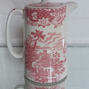 May include: A white ceramic pitcher with a lid, featuring a red and white Asian-inspired design. The pitcher has a handle and a spout. The design includes floral patterns, trees, and buildings.