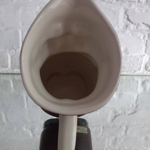 May include: A cream-colored ceramic pitcher with a long, curved spout and a handle. The pitcher has a wide opening and a dark interior. The handle is a light cream color, and the base is a dark brown color.