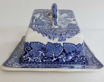 May include: A rectangular, white and blue ceramic butter dish with a lid and handle. The lid features a scenic landscape design, while the base is decorated with a leaf and floral pattern. The handle is a curved shape. The dish is designed for serving butter.