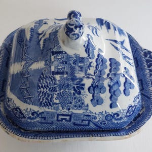 May include: Blue and white ceramic covered dish with a traditional Willow pattern. The dish has a rectangular base with rounded corners and a matching lid with a decorative handle. The design features scenes of a landscape with trees and buildings.