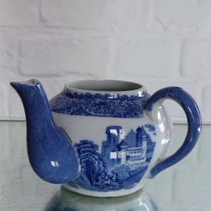 May include: A blue and white ceramic teapot with a landscape design. The teapot features a curved spout and handle, and the landscape scene includes buildings and trees. The teapot is sitting on a reflective surface.
