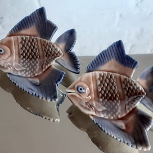 May include: Two ceramic fish figurines. The fish have a light peach body with brown and white details, and blue fins. The fish are displayed on a reflective surface. The fish are likely decorative items.