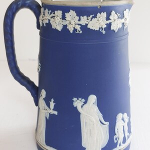 May include: A blue ceramic pitcher with a white relief design. The pitcher features a handle and a decorative border of grapevines. White relief figures are depicted on the body of the pitcher. The pitcher is a classic piece of home decor.