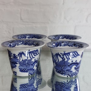 May include: Four white and blue ceramic planters with a classic Willow pattern. The planters have a flared rim and a detailed landscape design. The planters are reflected on a glass surface.