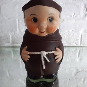 May include: A vintage ceramic figurine of a monk, featuring a brown robe and cowl, with a white rope belt. The figurine has a smiling face with rosy cheeks and a small red mouth. The base of the figurine is dark brown with a green trim.