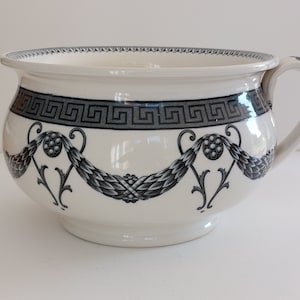 May include: White ceramic chamber pot with a handle and a decorative black and gray Greek key border. The pot features a garland design with floral accents, adding a touch of elegance to the piece.