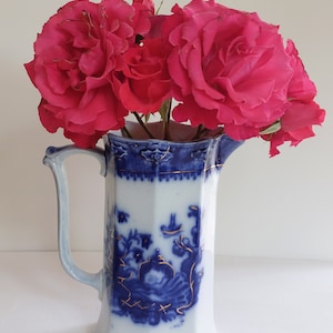 May include: A white and blue porcelain pitcher with a handle, filled with vibrant pink roses. The pitcher features a detailed blue floral design with gold accents. The roses are in full bloom, creating a striking contrast against the pitcher.