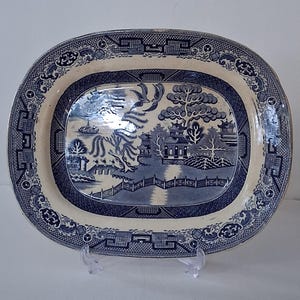 May include: Oval blue and white ceramic platter with a Willow pattern design. The platter features a central landscape scene with a bridge, trees, and buildings, surrounded by a decorative border. The platter is supported by a clear plastic stand.