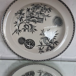 May include: Cream-colored decorative plate with black floral and geometric designs. The plate features a black border, a picture of a person reading, and other decorative elements. The plate is likely vintage.
