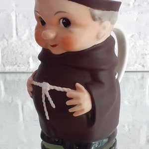 May include: A ceramic pitcher shaped like a cartoon monk, featuring a brown robe and a white handle. The monk has rosy cheeks, a brown cowl, and a white rope belt. The base is green and black.
