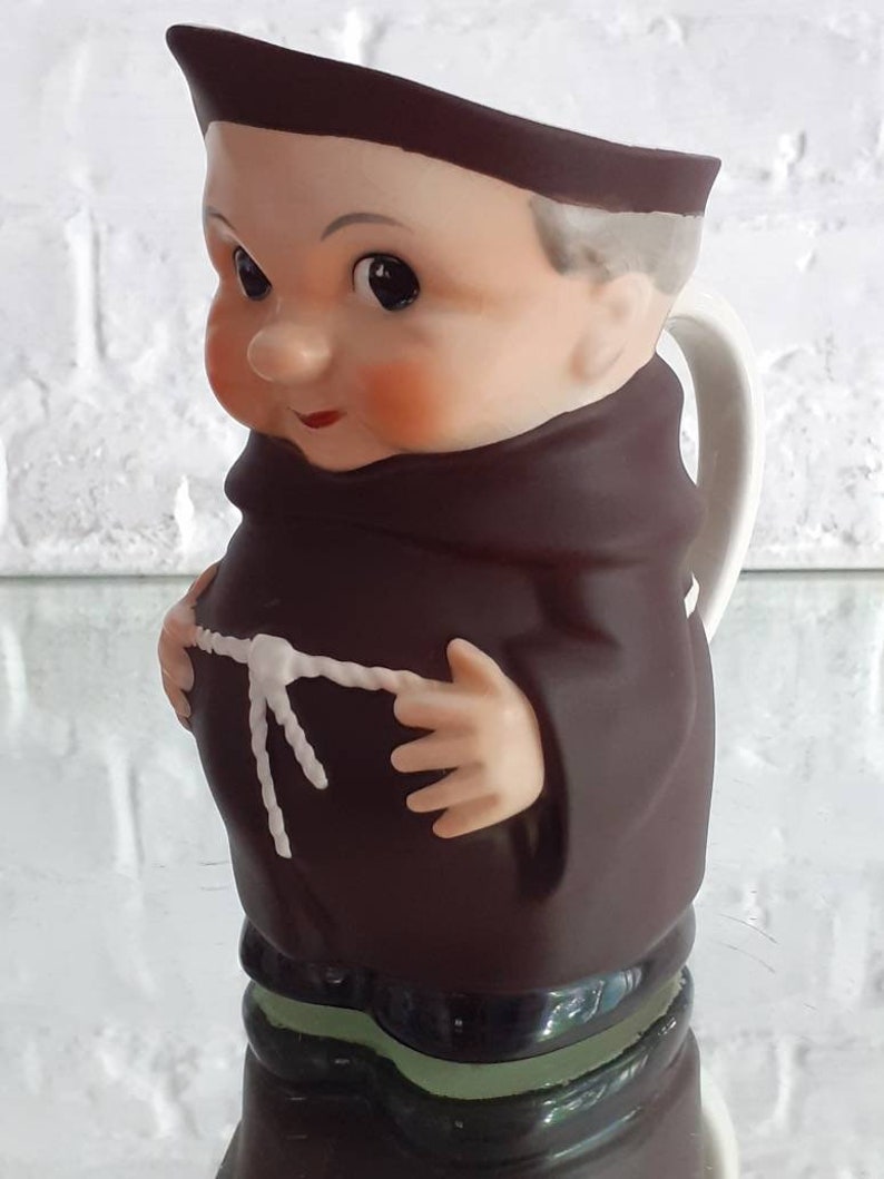 May include: A ceramic pitcher shaped like a cartoon monk, featuring a brown robe and a white handle. The monk has rosy cheeks, a brown cowl, and a white rope belt. The base is green and black.