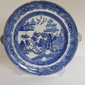 May include: Blue and white ceramic serving platter with a Willow pattern design. The platter features a detailed landscape scene with pagodas, bridges, and birds. It has a scalloped edge and two handles. A classic piece for serving.